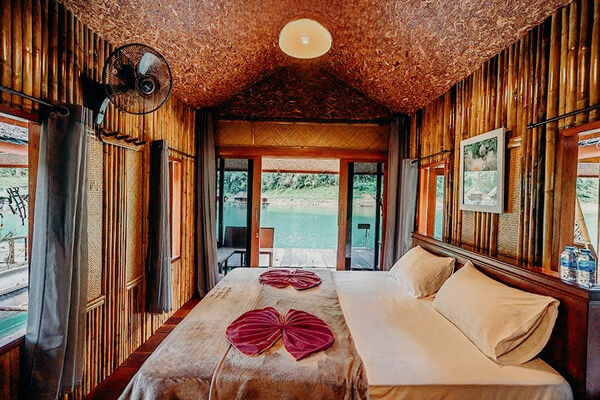 Wooden lodge bedroom with a double bed, folded towel on the duvet and ceiling fan, with open doors overlooking the lake in Thailand