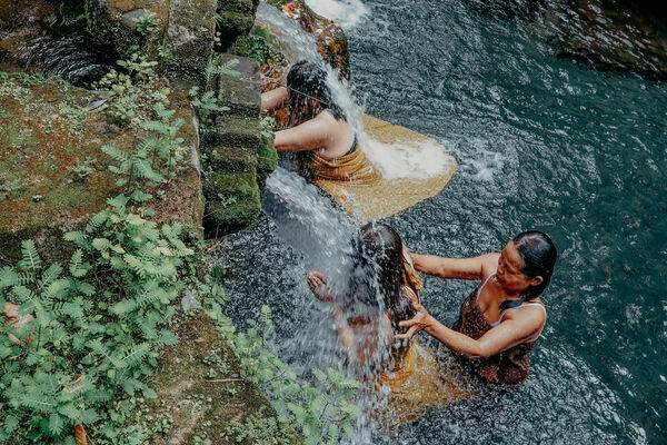 Participant performing Balinese melukat at a rocky river waterfall in Bali, hands lifted in cleansing water in a mossy gorge