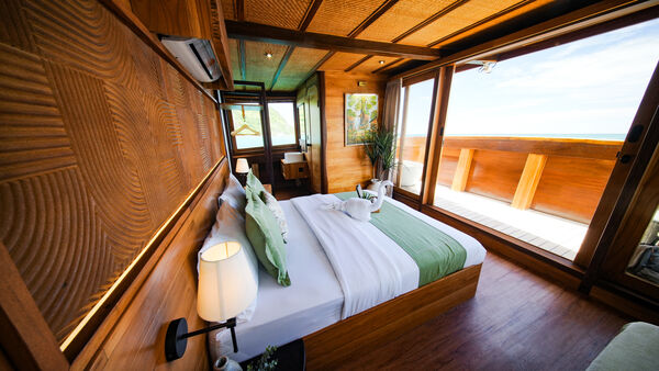 Elegant phinisi cabin with a made-up double bed and warm wood panelling, sunlight pouring through windows in Labuan Bajo