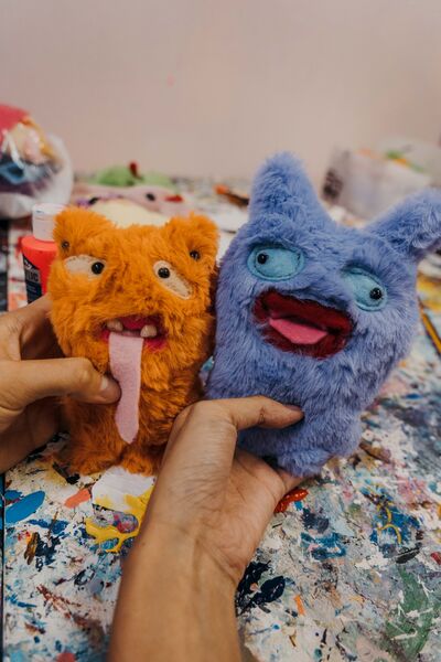 Hands holding orange and blue ugly-cute plushie keychains on a paint-splattered table at Avant-Garde Art Space workshops in Singapore