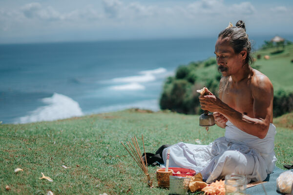 Jero Mangku Geni sits on a grassy cliff by the ocean with a ceremonial incense setup during spiritual healing in Bali
