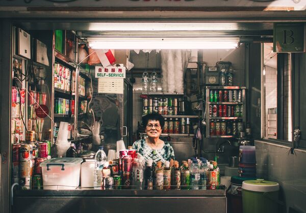 Chef in a Singapore hawker stall serving from behind a counter lined with jars and tins, with a “self-service” sign