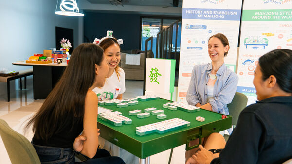 Coach guiding a group through mahjong at a green-topped table in Singapore, with classroom banners in the background