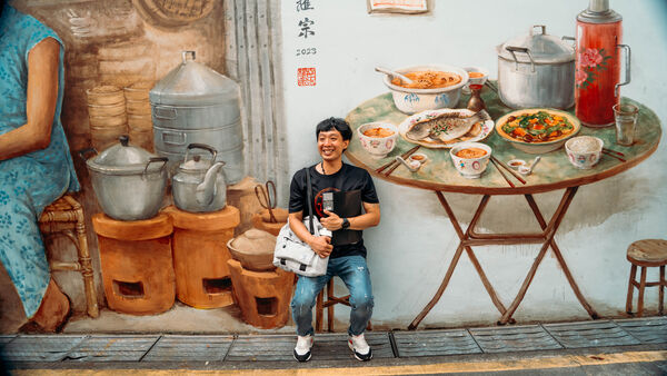 Tour participant holding a booklet and standing in front of a painted hawker stall with pots and noodle dishes in Singapore
