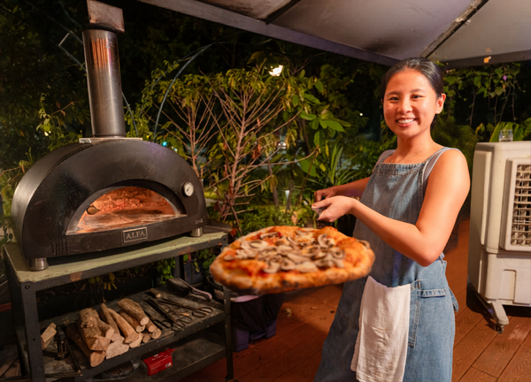 Woman in a denim apron holding a pizza fresh from a wood-fired oven in Singapore during hands-on workshops in Singapore