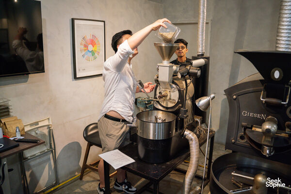 Participant loading coffee into a roaster grinder at Compound Coffee Co. in Singapore, inside the private roasting workshop room