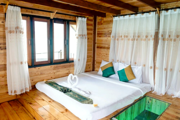 Wooden rafthouse bedroom with a neatly made white bed, green pillows and sheer curtains, overlooking Khao Sok National Park