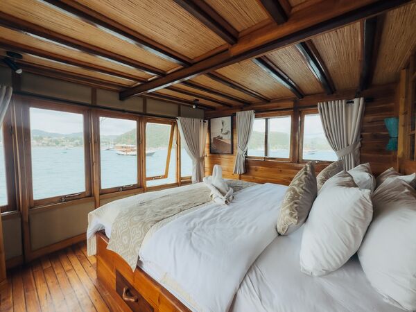 Phinisi suite cabin with a large double bed and folded towels, wooden windows framing the water, in Labuan Bajo