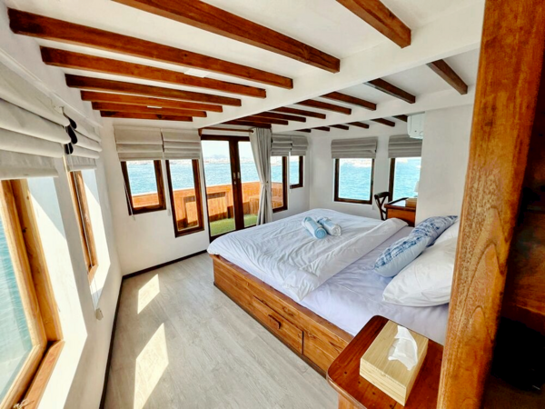 Wood-beamed phinisi cabin with a low double bed, neatly rolled towels, and large windows framing the sea, in Labuan Bajo