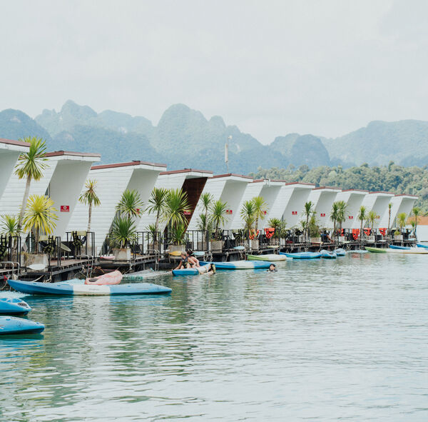 Cabin row beside a calm lake with moored long-tail boats and karst mountain views, at Cheow Lan Lake