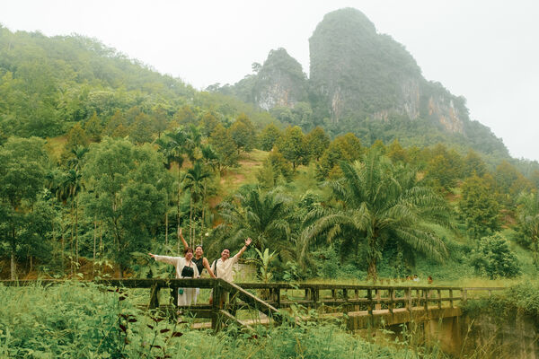 People smiling on a wooden riverboat in Thailand, with lush jungle palms and steep green hills rising in misty weather