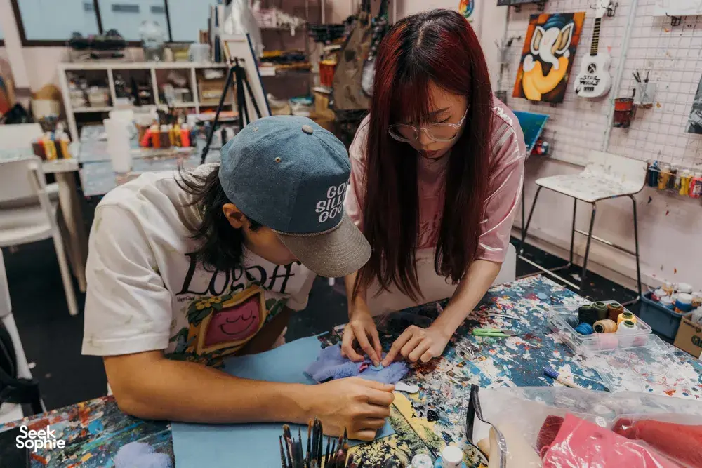 Two participants stitching an ugly-cute plush keychain on a shared table in a Singapore art studio, surrounded by tools