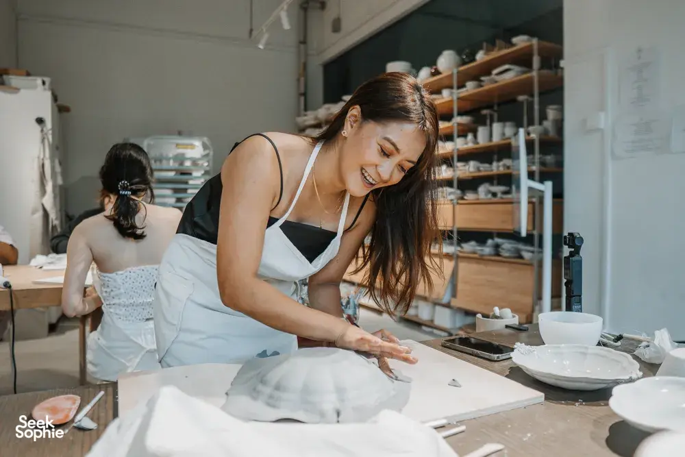 Singapore pottery workshop: instructor shaping a ceramic plate on a workbench with tools, as another participant works in the back