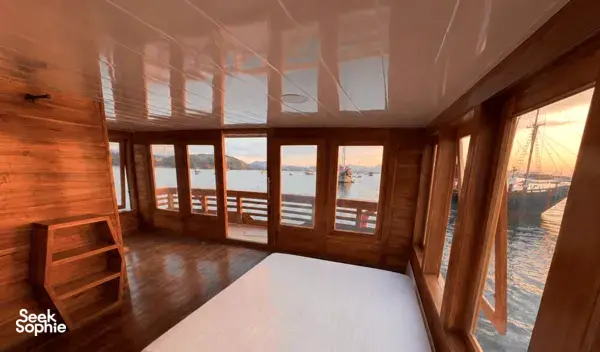 Wooden phinisi lounge with windowed walls and a view over the Komodo waters on a shared 3D2N cruise from Labuan Bajo