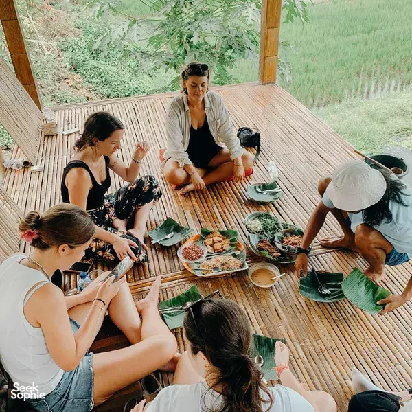 People sit around a wooden deck table sharing a meal in Lombok during the 4D3N loop, with lush hills in the background