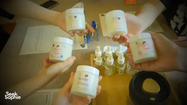 Hands raised holding labelled coco-soy candle jars over a table of oils and wicks during a Singapore candle workshop