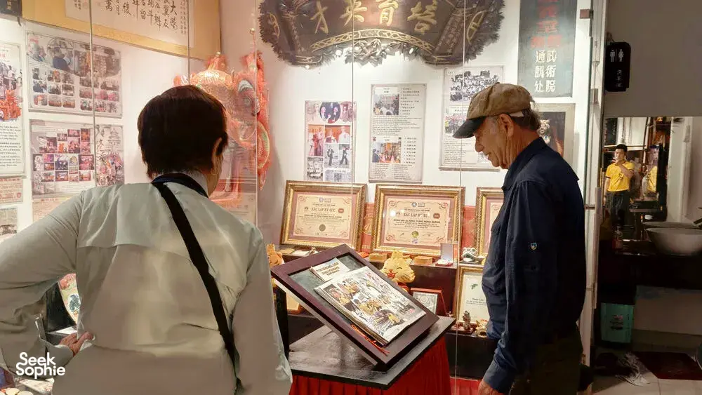 Ho Chi Minh City cultural display: an older host and visitor browse lion dance documents, photos and framed certificates