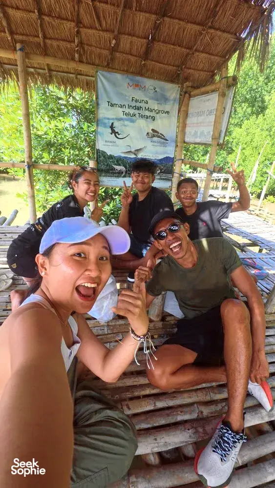 Sekumpulan pengembara tersenyum di ruang tempat duduk kayu berbumbung rumbia di sebelah papan tanda ilustrasi bakau di Labuan Bajo