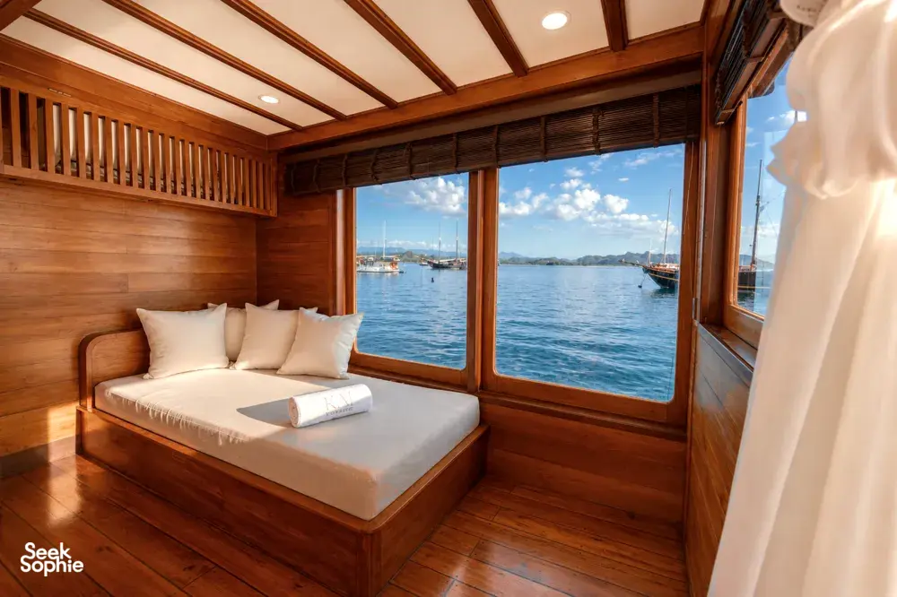 Wooden phinisi daybed with white cushions and pillows, set beside large windows with blue sea and boats in Labuan Bajo