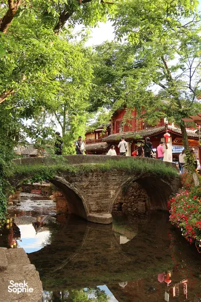 People walking past an arched stone bridge over a canal in Shuhe Ancient Town, Lijiang, with lush trees and lanterns nearby.