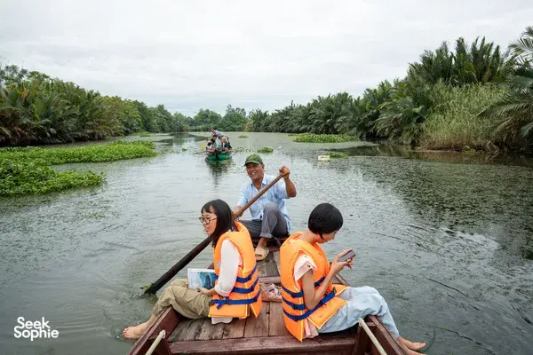 Life-jacketed travellers ride a wooden boat along a calm, palm-lined river in Hoi An on a conservation-led journey