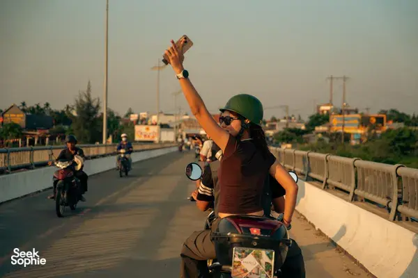 Helmed Vespa rider on a bridge road in Hoi An, arm raised as the guided tour rides out at golden hour