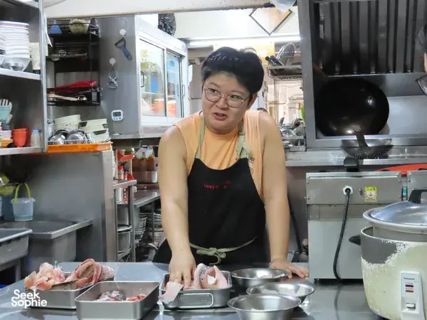 Female hawker preparing Cantonese fish at a Singapore street-food stall, with diners nearby observing from behind