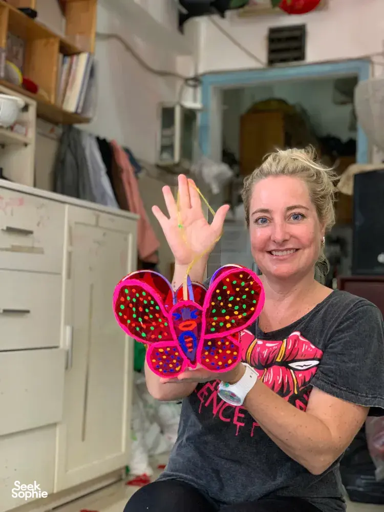 Smiling guest holding colourful butterfly-shaped crafts on a shelf-lined workshop table in Ho Chi Minh City, Viet Awesome Tours