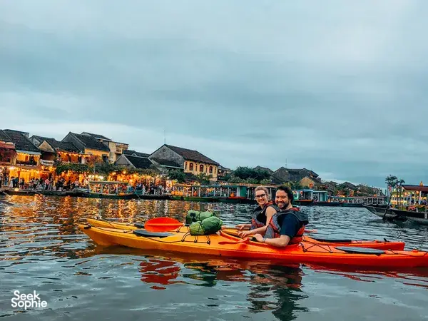 Two travellers smile aboard an orange kayak on a Hoi An Kayak Tour, passing lit riverside buildings in Hoi An