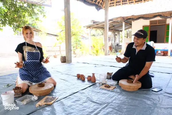 Two people sit with artisans at a pottery village craft session, shaping small clay pieces on mats in Yogyakarta