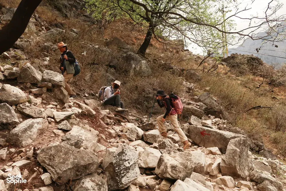 Several hikers cross a steep rocky hillside on the Tiger Leaping Gorge high trail in Lijiang, with sparse trees along the track