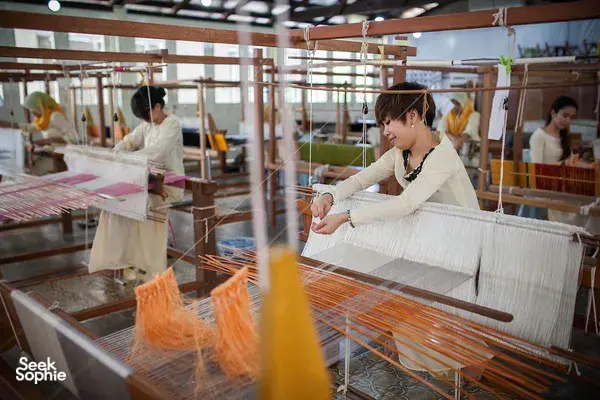 Woman weaving on a handloom inside a bright Tanoti Crafts atelier in Kuching, with more looms and weavers in rows