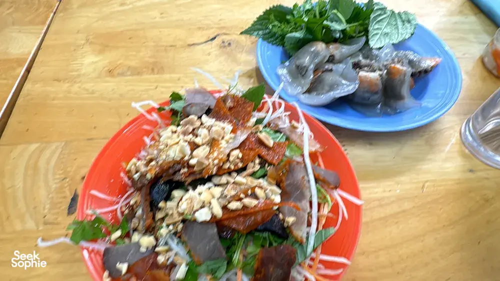 Assorted Hanoi street-food—grilled meats with herbs—served on a red plate on a wooden table for a Zero‑Tourist food adventure