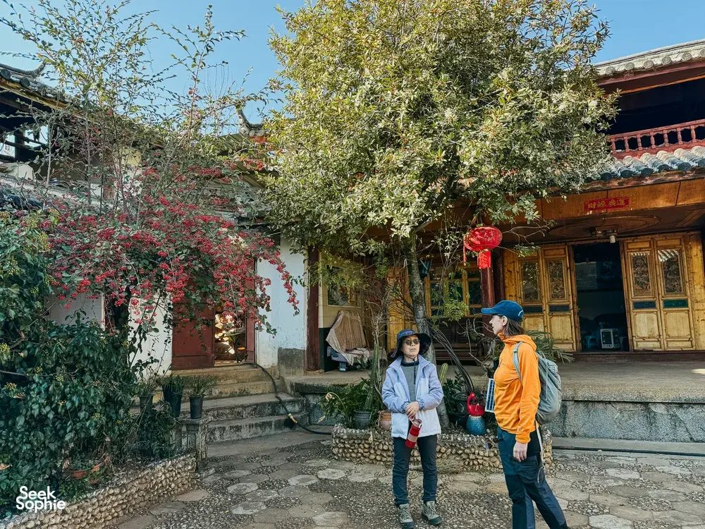 Two travellers walking a stone courtyard in Lijiang, passing flowering trees beside a traditional wooden door during the trek