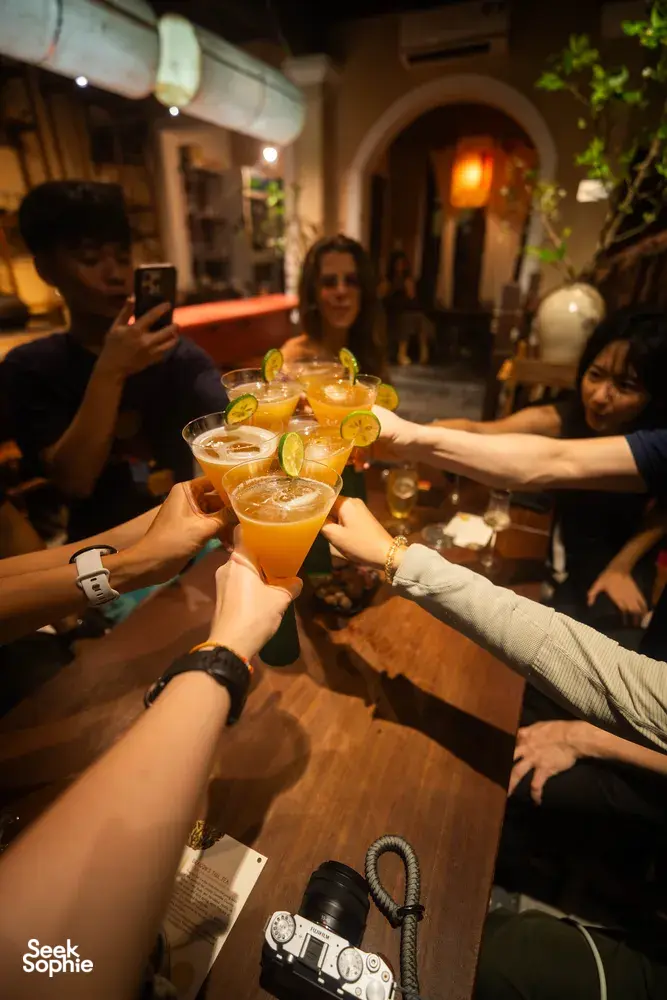 在会安鸡尾酒之旅中，朋友们在灯笼映照的庭院里举起橙色鸡尾酒杯碰杯