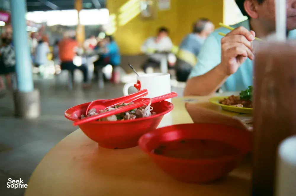 Red hawker bowls on a dining table in Singapore, with diners’ hands enjoying noodles and side dishes together
