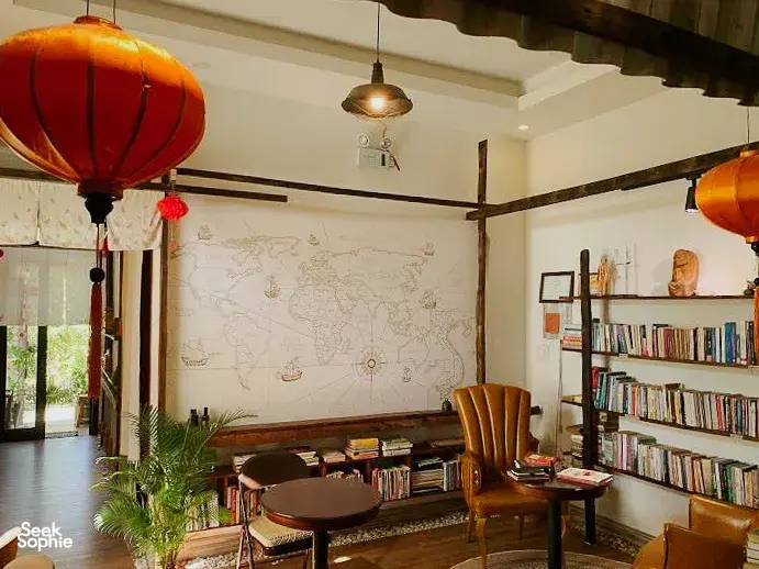 Area lounge di Chan Dat Living Room di Hoi An dengan kursi, rak buku, dinding peta dunia, dan lampion oranye gantung