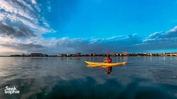 Aerial view of a kayaker on clear water in Hoi An, reflecting the evening skyline as the paddle moves forward