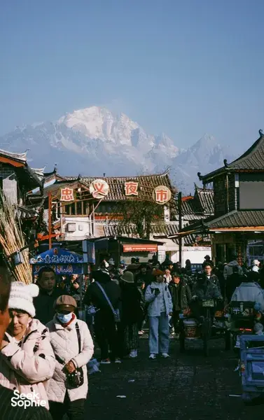 Evening street market in Lijiang with lantern-lit shopfronts, shoppers bundled up, and a snow-capped mountain towering behind