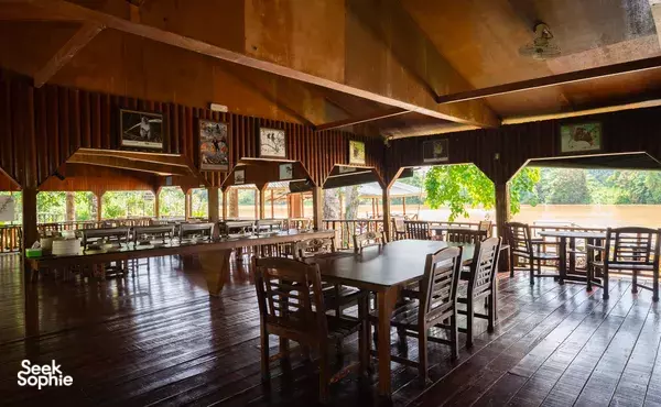 Open-air wooden dining hall with rows of chairs and long tables, framed wall photos, and leafy views through windows