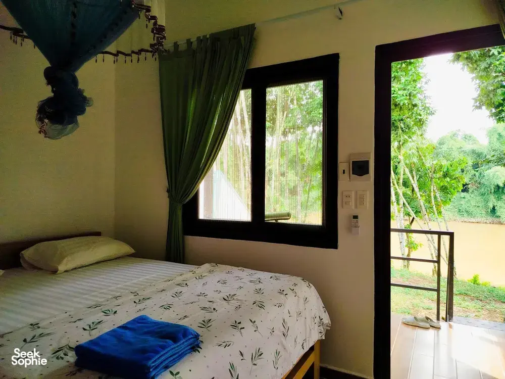 Green Hope Lodge bedroom corner with patterned duvet and green curtains, looking out from the terrace to rainforest greenery at Cat Tien