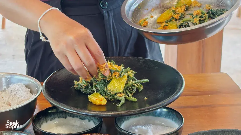 Hands arranging vegan Manggarai food on a black skillet plate during a hands-on cooking class in Labuan Bajo