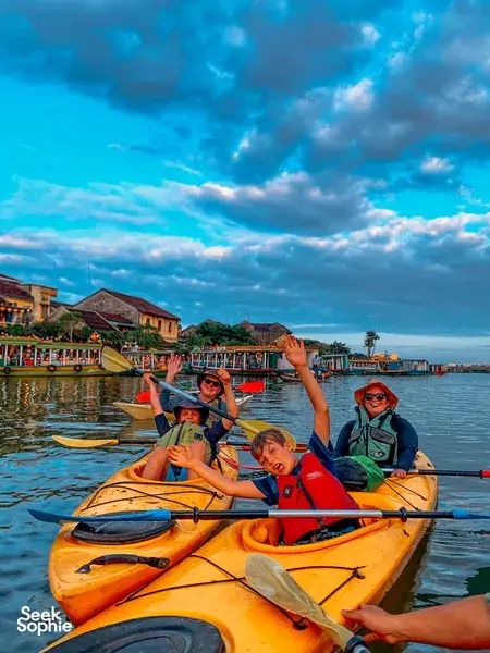 Group in orange and yellow kayaks smiling and raising paddles in Hoi An harbour, with riverfront houses behind them