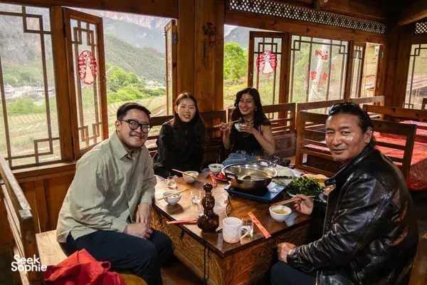 Group sharing a meal inside a traditional dining room in Shangri-La, with large windows framing a green mountain valley