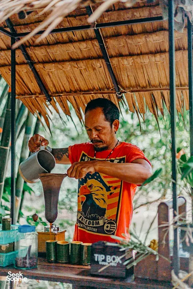 Community stall worker pouring from a large kettle under a palm roof in Khao Lak for a conservation-focused tour