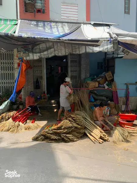 Straw brooms and bundles on the ground outside a Saigon craft alley workshop, where an artisan tends materials by the door
