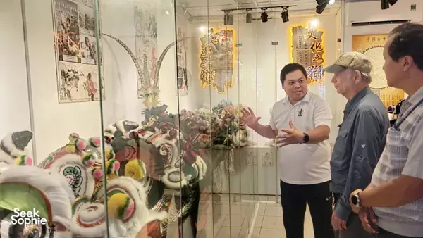 Museum gallery scene in Ho Chi Minh City Chinatown, where a guide gestures at lion dance costumes and visitors listen to the history