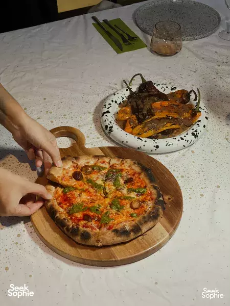 Hands serving a stone-baked pizza from a wooden board, with plated mains and drinks on a table in Singapore for private dining Singapore