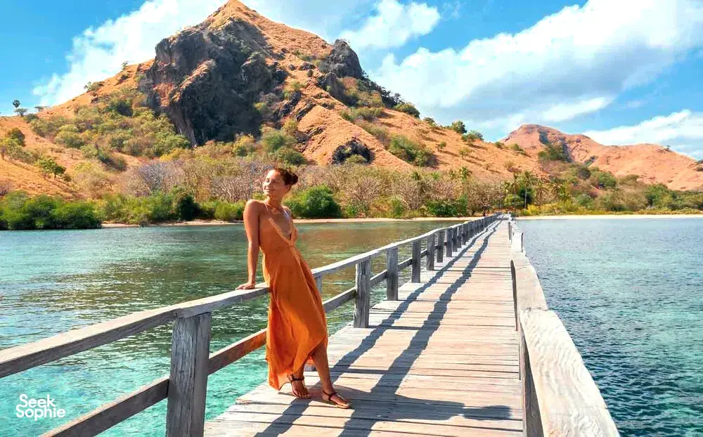 Traveller in a rust-coloured dress walking along a wooden pier over clear water with rugged hills in the distance, in Labuan Bajo