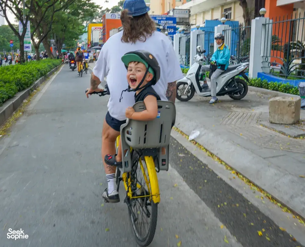在胡志明市一条林荫街道上骑行时,一名戴着头盔、坐在自行车座椅上的孩子开心地笑着。